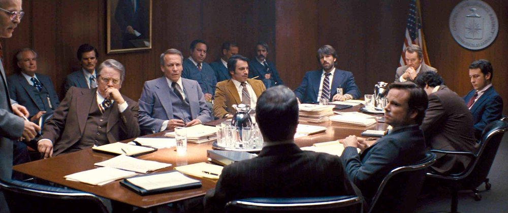 Photo of a CIA strategy meeting, featuring Zeljko Ivanek (standing), with Chris Messina, Ben Affleck, Bryan Cranston, and others, seated at the opposite end of the table.